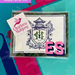May include: A clear plastic sleeve containing a 2026 International Mahjong card. The card features a purple pagoda crest with the letters CM, surrounded by floral details and a green plaid border. A white sticker reads Charlotte Mahjong with a pink bird logo.
