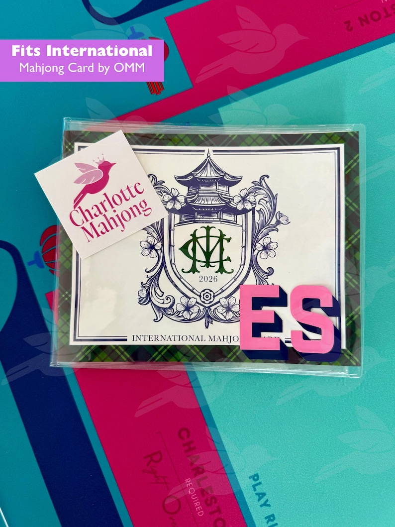 May include: A clear plastic sleeve containing a 2026 International Mahjong card. The card features a purple pagoda crest with the letters CM, surrounded by floral details and a green plaid border. A white sticker reads Charlotte Mahjong with a pink bird logo.