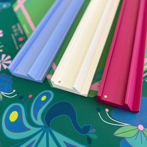 May include: Three colorful wooden picture ledges in light blue, white, and magenta. The ledges are displayed on a green surface with a peacock and floral pattern. The ledges are designed to display photos or artwork.