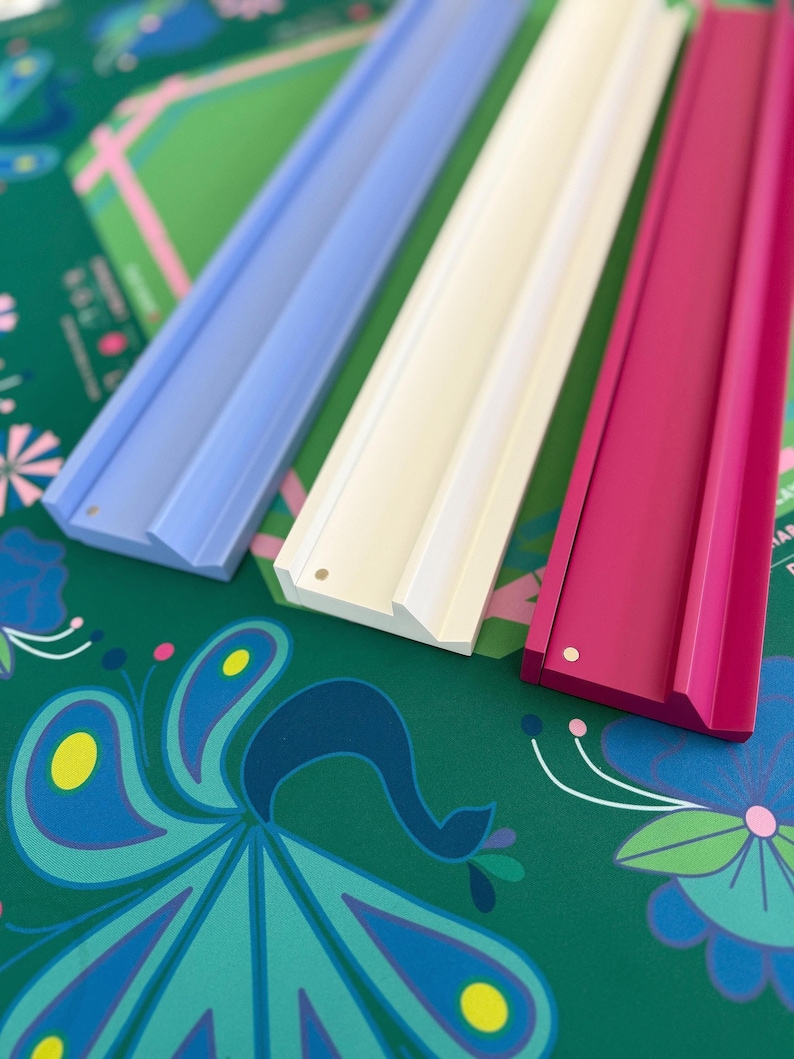 May include: Three colorful wooden picture ledges in light blue, white, and magenta. The ledges are displayed on a green surface with a peacock and floral pattern. The ledges are designed to display photos or artwork.
