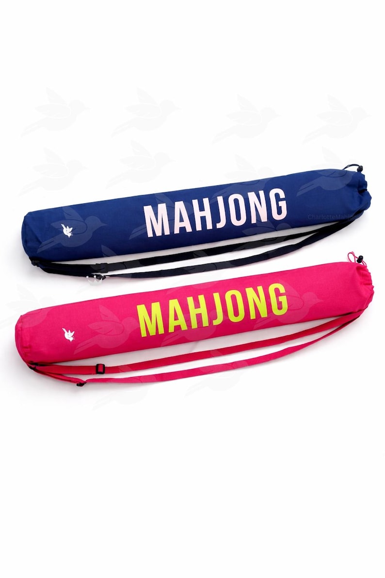 Puede incluir: Dos estuches cil&iacute;ndricos para fichas de Mahjong. Uno es azul marino con texto blanco y una correa negra. El otro es rosa intenso con texto amarillo-verdoso y una correa rosa. Ambos estuches tienen un patr&oacute;n de p&aacute;jaros.