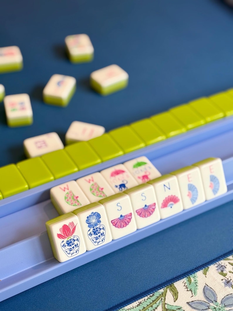May include: A close-up of a Mahjong set with white tiles featuring colorful illustrations and letters. The tiles are arranged in a blue tray with green and blue accents. The background is a solid blue surface.