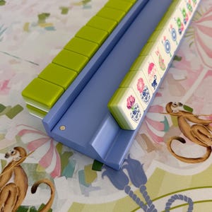 May include: A Mahjong set with green and white tiles in a light blue tray. The tiles feature colorful illustrations. The set is on a patterned surface with monkeys and floral designs.