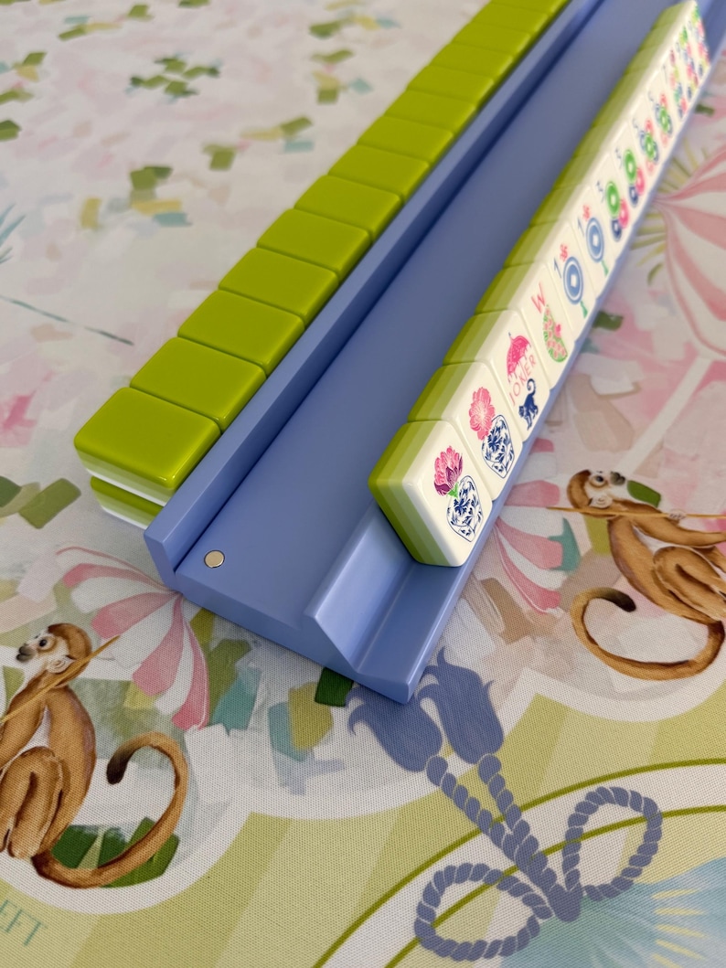 May include: A Mahjong set with green and white tiles in a light blue tray. The tiles feature colorful illustrations. The set is on a patterned surface with monkeys and floral designs.