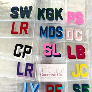 May include: A collection of clear plastic pouches, each containing a pair of block-letter initials in various colors including navy, green, pink, red, black, purple, light blue, and yellow. A white card with the text A Polished Greeting Co. is included.