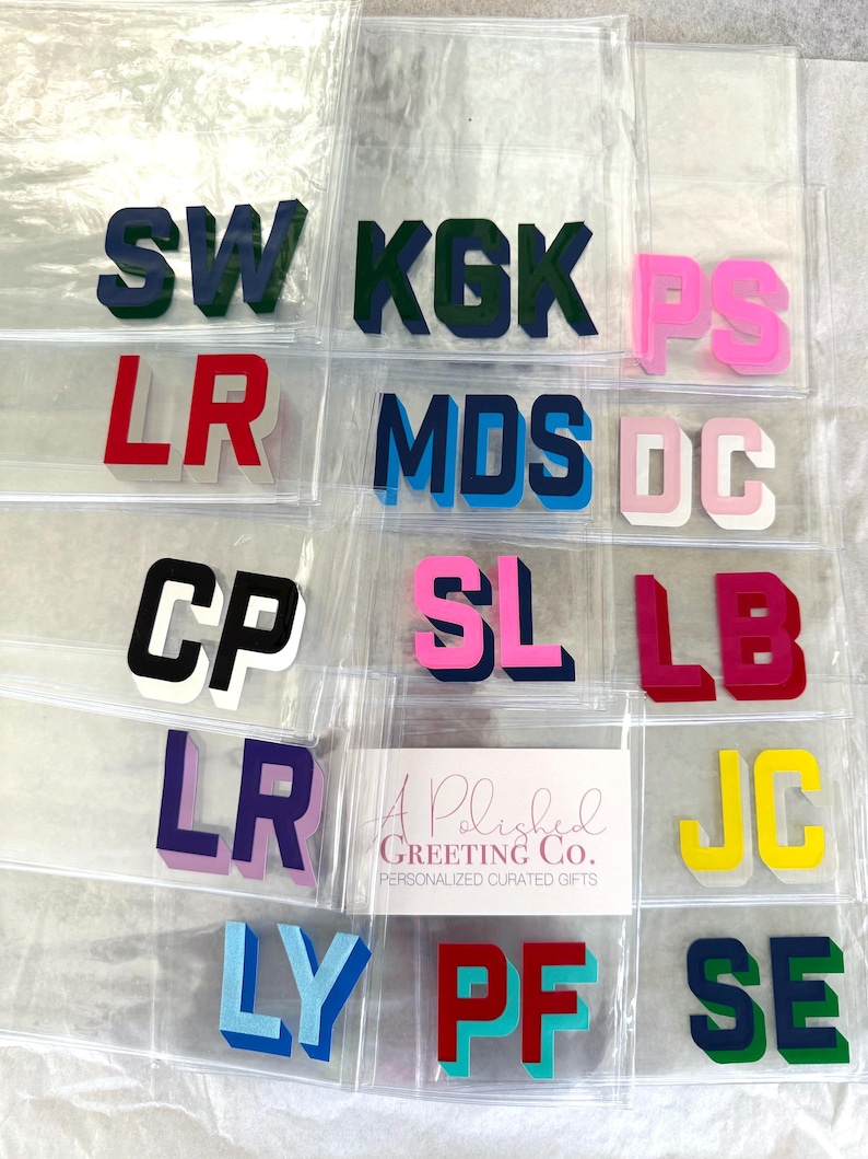May include: A collection of clear plastic pouches, each containing a pair of block-letter initials in various colors including navy, green, pink, red, black, purple, light blue, and yellow. A white card with the text A Polished Greeting Co. is included.