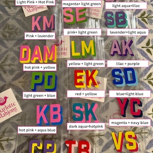 May include: A collection of two-letter acrylic mahjong tiles in various color combinations, including pink, yellow, blue, green, and purple. Each tile set is packaged in a clear plastic sleeve with a Charlotte Mahjong brand label.