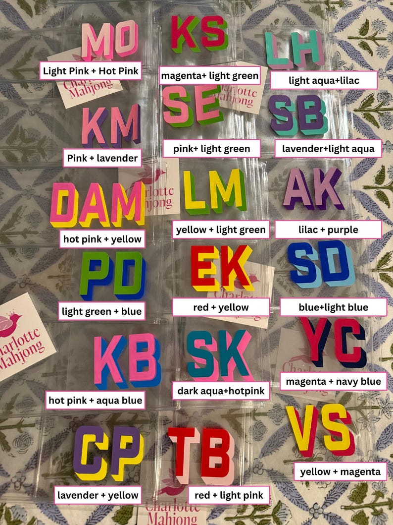 May include: A collection of two-letter acrylic mahjong tiles in various color combinations, including pink, yellow, blue, green, and purple. Each tile set is packaged in a clear plastic sleeve with a Charlotte Mahjong brand label.