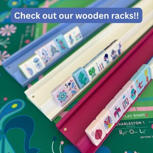 May include: Three wooden Mahjong racks in light blue, white, and magenta, each holding a row of white tiles with colorful floral and numerical designs. The text "Check out our wooden racks!!" is displayed in a blue banner at the top of the image.