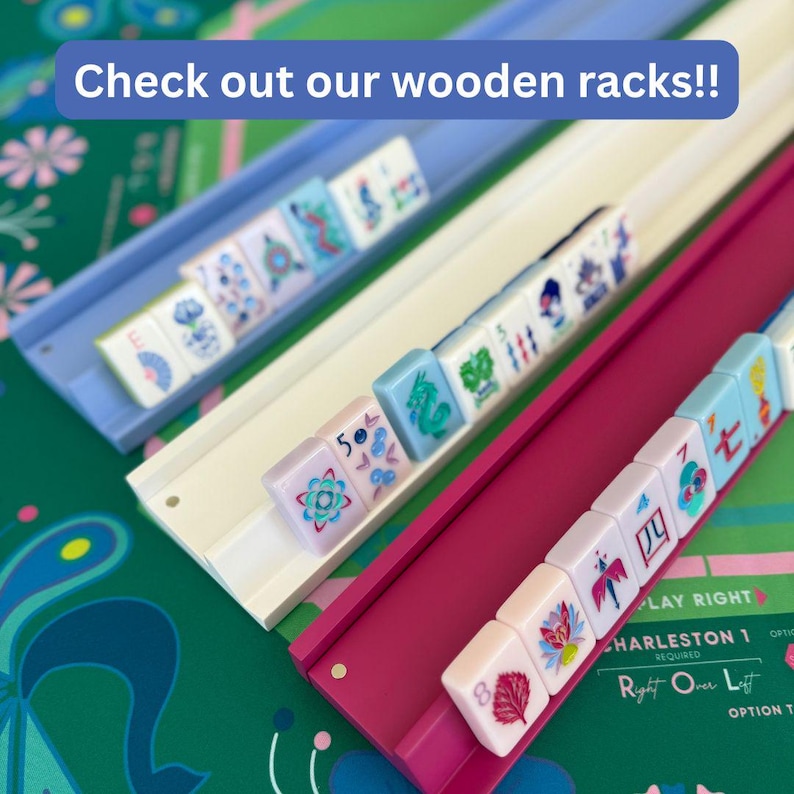 May include: Three wooden Mahjong racks in light blue, white, and magenta, each holding a row of white tiles with colorful floral and numerical designs. The text "Check out our wooden racks!!" is displayed in a blue banner at the top of the image.