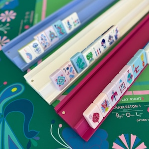 May include: Three colorful Mahjong tile racks in blue, white, and magenta, each holding a row of tiles. The tiles feature various symbols and designs. The background is a green surface with floral patterns and text that reads "PLAY RIGHT CHARLESTON 1".