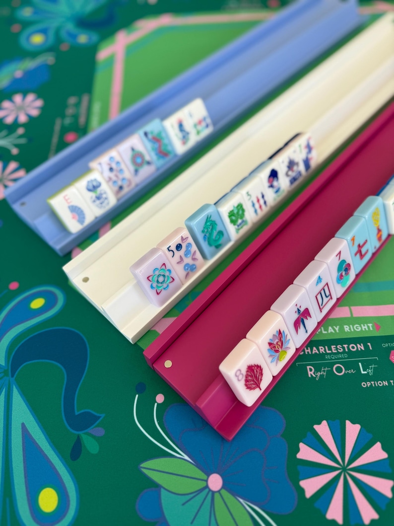 May include: Three colorful Mahjong tile racks in blue, white, and magenta, each holding a row of tiles. The tiles feature various symbols and designs. The background is a green surface with floral patterns and text that reads "PLAY RIGHT CHARLESTON 1".