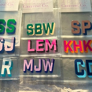 May include: A collection of acrylic monogram letter sets in various color combinations, including green, pink, orange, blue, and lavender. Each set features block-style letters with a 3D shadow effect, individually packaged in clear plastic sleeves.