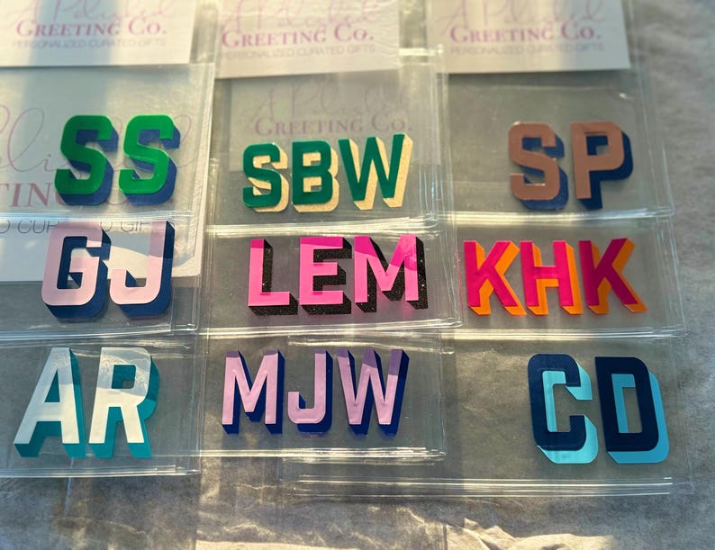May include: A collection of acrylic monogram letter sets in various color combinations, including green, pink, orange, blue, and lavender. Each set features block-style letters with a 3D shadow effect, individually packaged in clear plastic sleeves.