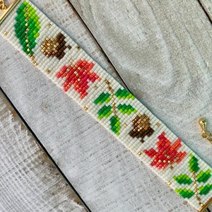May include: A white beaded bracelet with a repeating pattern of green leaves, red and orange maple leaves, and brown acorns. The bracelet has gold-toned clasps.