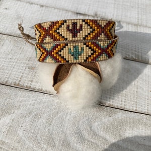May include: Two beaded bracelets with a geometric pattern in shades of brown, yellow, blue, and white. The bracelets are stacked on top of each other and are tied together with a brown leather cord.