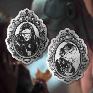 Puede incluir: Dos pegatinas en blanco y negro con retratos en marcos ovalados ornamentados. Una representa a una persona con pelo largo y expresión severa, la otra a una persona con el pelo recogido y la cabeza baja. Las pegatinas tienen una estética vintage.