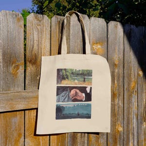 May include: Beige tote bag with three rectangular images. The top image shows a person fishing by a lake. The middle image shows a person's hand. The bottom image shows a person walking in a field. The bag hangs on a wooden fence.