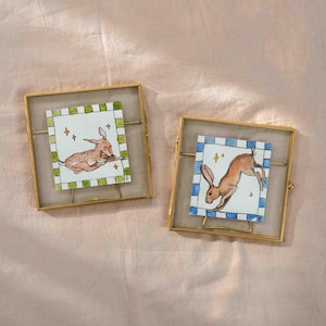 May include: Two square framed art pieces with watercolor illustrations of rabbits. One shows a brown rabbit lying down, the other a jumping rabbit. Each is set against a white background with a checkered border and gold frames.