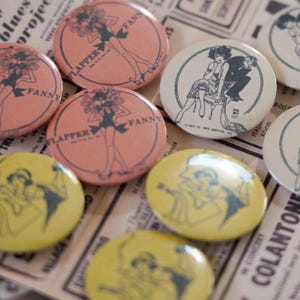 Peut inclure: Plusieurs badges vintage avec des illustrations de flappers. Les badges sont de différentes couleurs, notamment rose, jaune et blanc cassé, avec des graphiques noirs et gris. Certains badges affichent le texte "FLAPPER FANNY".