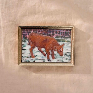 May include: An oil painting in a gold frame depicts a brown cow in a snowy field. The cow is the focal point, with a white marking on its forehead. The background shows a brick wall.