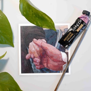 Puede incluir: Un primer plano de una pintura de dos manos entrelazadas. La pintura est&aacute; en una tarjeta blanca con un tubo de pintura gouache rosa y un pincel encima.