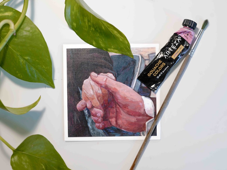 Puede incluir: Un primer plano de una pintura de dos manos entrelazadas. La pintura est&aacute; en una tarjeta blanca con un tubo de pintura gouache rosa y un pincel encima.