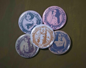 Könnte beinhalten: Fünf runde Ansteckbuttons mit Porträts von Personen, die von verzierten, dekorativen Rahmen umgeben sind. Die Buttons zeigen verschiedene Farbkombinationen, darunter Helllila, zartes Rosa, Weiß und Beige, mit kontrastierenden Illustrationen in dunkler Tinte.