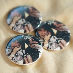 Op de afbeelding: Drie ronde buttons met een fotoprint van twee mensen met donker haar. Het ontwerp bevat witte sterpictogrammen en wervelende lijngraphics die over de donkere, sfeervolle afbeelding zijn geplaatst, met een glanzende, reflecterende afwerking.