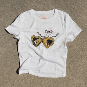 May include: A white short-sleeved t-shirt featuring a graphic print of a gold-colored heart-shaped locket that is open. The locket displays a profile illustration of a person on each side and is tied with a decorative red and white ribbon.