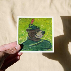 May include: A hand-painted illustration of a brown bear wearing a green hat with a red feather. The bear is looking to the right and has a friendly expression. The background is a green field with white dots.