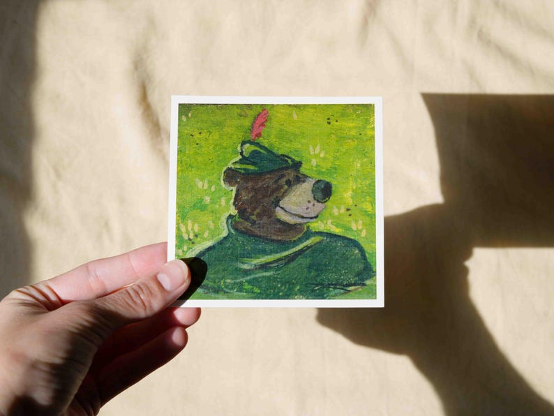 May include: A hand-painted illustration of a brown bear wearing a green hat with a red feather. The bear is looking to the right and has a friendly expression. The background is a green field with white dots.