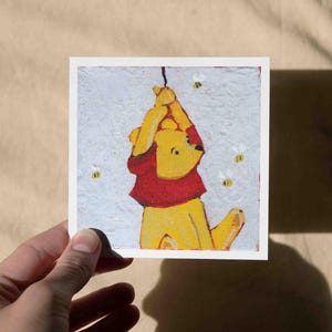 May include: A square art print featuring a yellow bear in a red shirt, hanging from a string. The bear is surrounded by three small bees. The artwork has a textured, painted appearance.