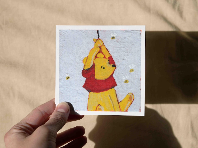May include: A square art print featuring a yellow bear in a red shirt, hanging from a string. The bear is surrounded by three small bees. The artwork has a textured, painted appearance.
