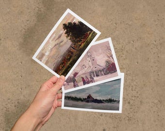 May include: Three rectangular postcards featuring printed landscape and architectural illustrations. One shows a sunset over a field with a large tree, another depicts a church building with figures in front, and the third shows a figure on a beach.