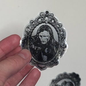 May include: A silver sticker featuring a black and white portrait within an ornate frame. The portrait depicts a person with long hair and a somber expression. The sticker has a glossy finish and is held by a hand.