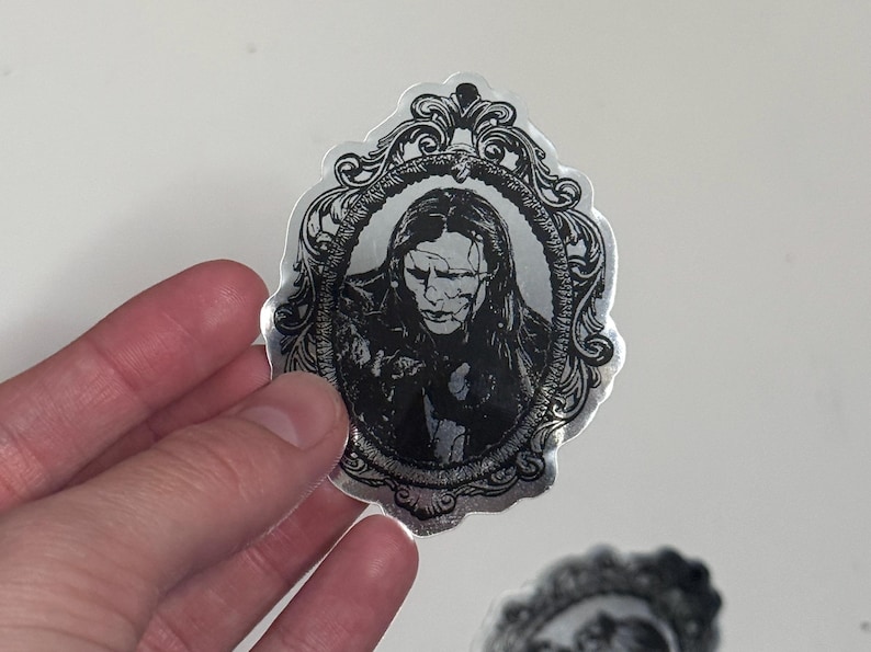 May include: A silver sticker featuring a black and white portrait within an ornate frame. The portrait depicts a person with long hair and a somber expression. The sticker has a glossy finish and is held by a hand.