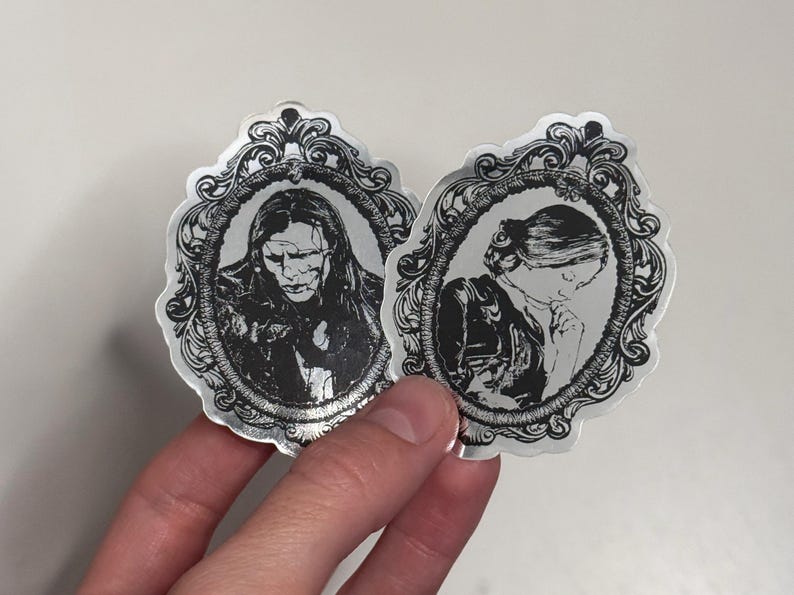 May include: Two silver stickers featuring black and white portraits within ornate frames. One depicts a person with long hair and a somber expression, the other a person with a blindfold. The stickers have a vintage, gothic aesthetic.