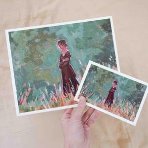 May include: Two art prints depicting a woman in a long, dark dress in a field. The larger print is held in a hand, with a smaller version visible. The artwork has a painterly style with green and brown tones.