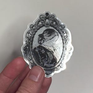May include: Silver sticker featuring a black and white portrait of a person in an ornate oval frame. The portrait shows a person with their hair up and wearing a high-collared garment. The sticker has a decorative, vintage aesthetic.