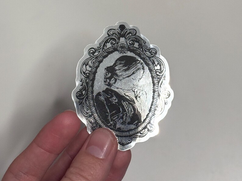May include: Silver sticker featuring a black and white portrait of a person in an ornate oval frame. The portrait shows a person with their hair up and wearing a high-collared garment. The sticker has a decorative, vintage aesthetic.