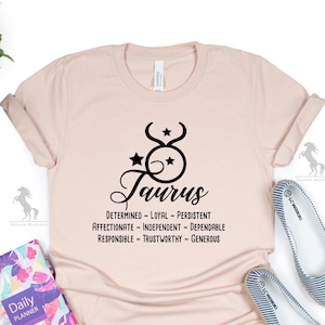 May include: A light pink t-shirt with a black Taurus zodiac symbol and the text "Taurus" in a cursive font. The text "Determined - Loyal - Persistent - Affectionate - Independent - Dependable - Responsible - Trustworthy - Generous" is printed below the symbol.