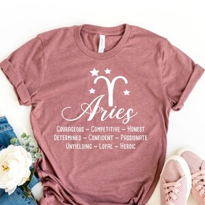 Może przedstawiać: R&oacute;żowy T-shirt z białym nadrukiem przedstawiającym znak zodiaku Baran i słowo "Baran" w ozdobnym, pochylonym kroju pisma. Pod nadrukiem znajduje się tekst "COURAGEOUS - COMPETITIVE - HONEST DETERMINED - CONFIDENT - PASSIONATE UNYIELDING - LOYAL - HEROIC".