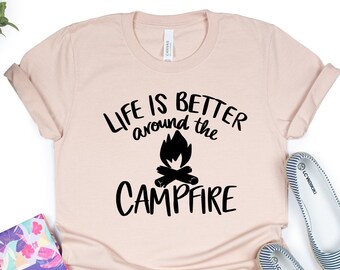 May include: A light pink t-shirt with the text "Life is better around the campfire" in black lettering. A black silhouette of a campfire with flames is centered between the words "around" and "the".