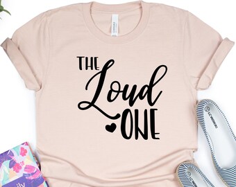 Könnte beinhalten: Ein hellrosa T-Shirt mit dem Text "The Loud One" in schwarzer Schrift. Das Shirt liegt auf einem weißen Hintergrund mit einem Blumen-Planer und einem Paar blau-weiß gestreiften Schuhen.