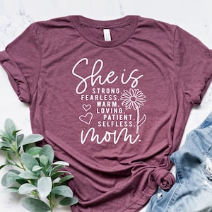 Może przedstawiać: Koszulka w kolorze bordowym z napisem "She is strong, fearless, warm, loving, patient, selfless, mom." w białym kolorze. Tekst jest otoczony małym kwiatkiem i sercami.