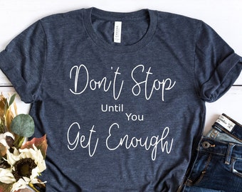 May include: A navy blue t-shirt with white text that reads "Don't Stop Until You Get Enough". The shirt is laying on a white wooden surface with a pair of brown sandals and blue jeans.