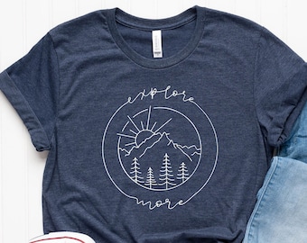 May include: A navy blue t-shirt featuring a white graphic of a mountain range, sun, and trees in a circle with the text "explore more". The shirt is paired with light blue jeans and white trainers.