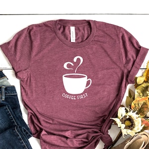 May include: A maroon t-shirt featuring a white graphic of a coffee cup with steam rising from it and the text "Coffee First". The shirt is laid out on a white wooden surface with a pair of blue jeans, a small white pumpkin, and a bouquet of dried flowers.