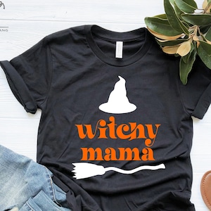 May include: Black t-shirt with a white witch hat and a broom graphic. The text "witchy mama" is printed in orange on the shirt.